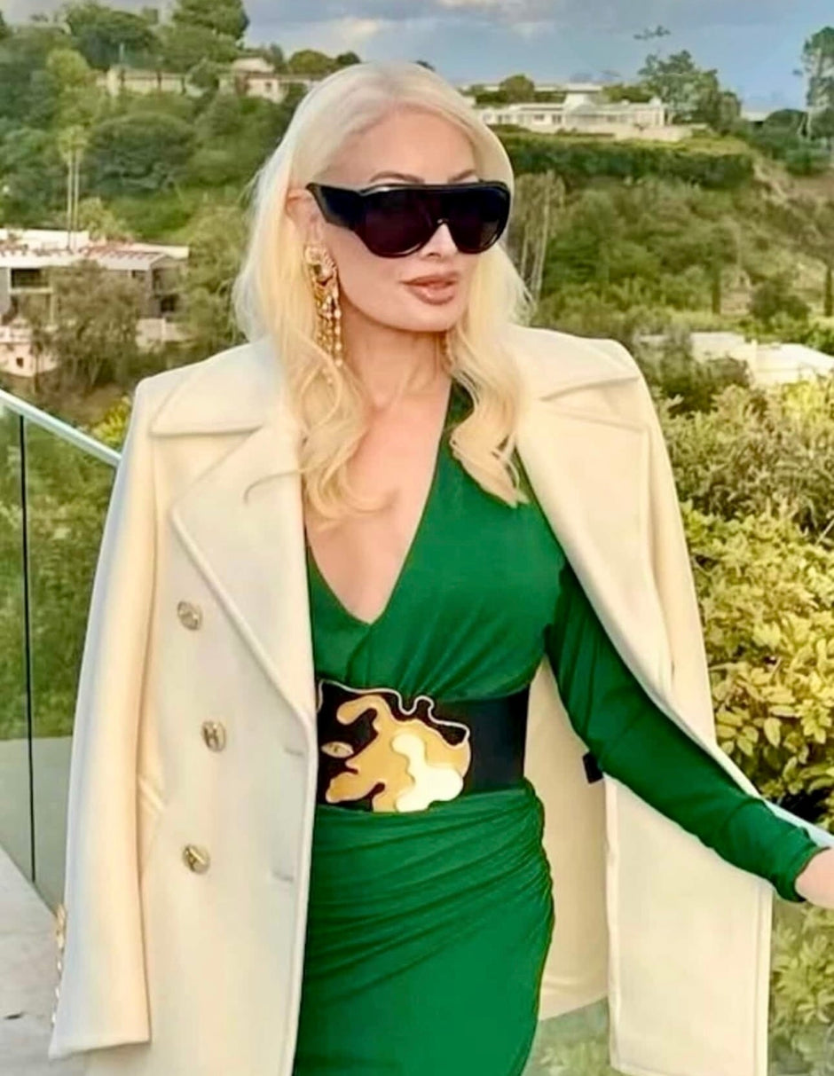 Woman in a green dress with a beige coat and sunglasses outdoors